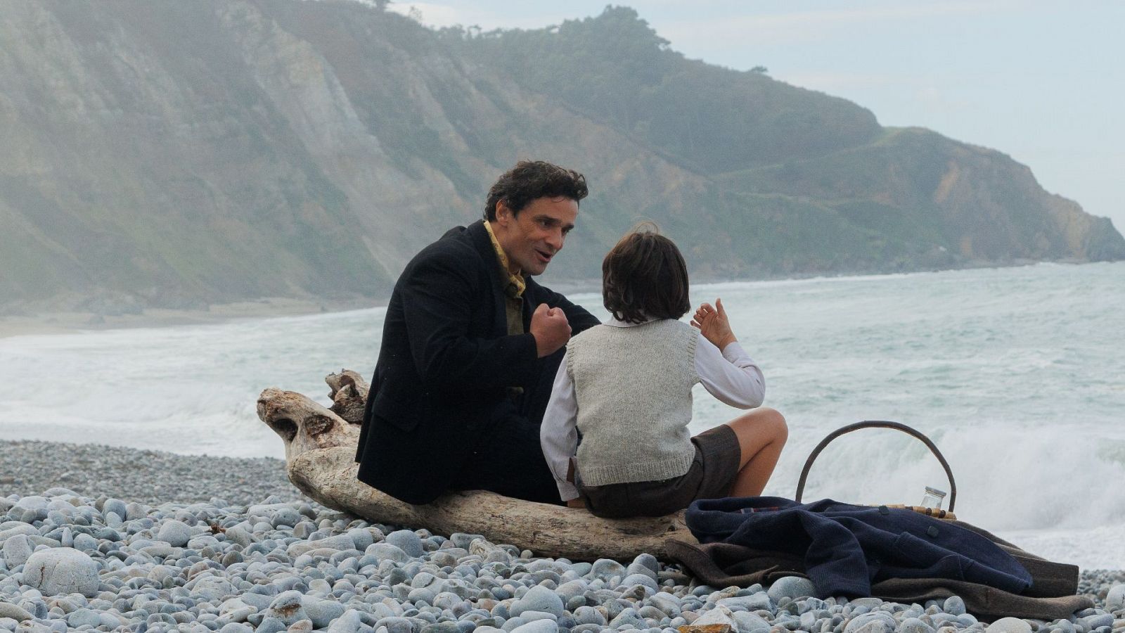 Abuelo y nieto conversan en una playa rocosa, junto a una cesta y ropa. El niño viste chaleco claro y el adulto ropa oscura. Escena tranquila.