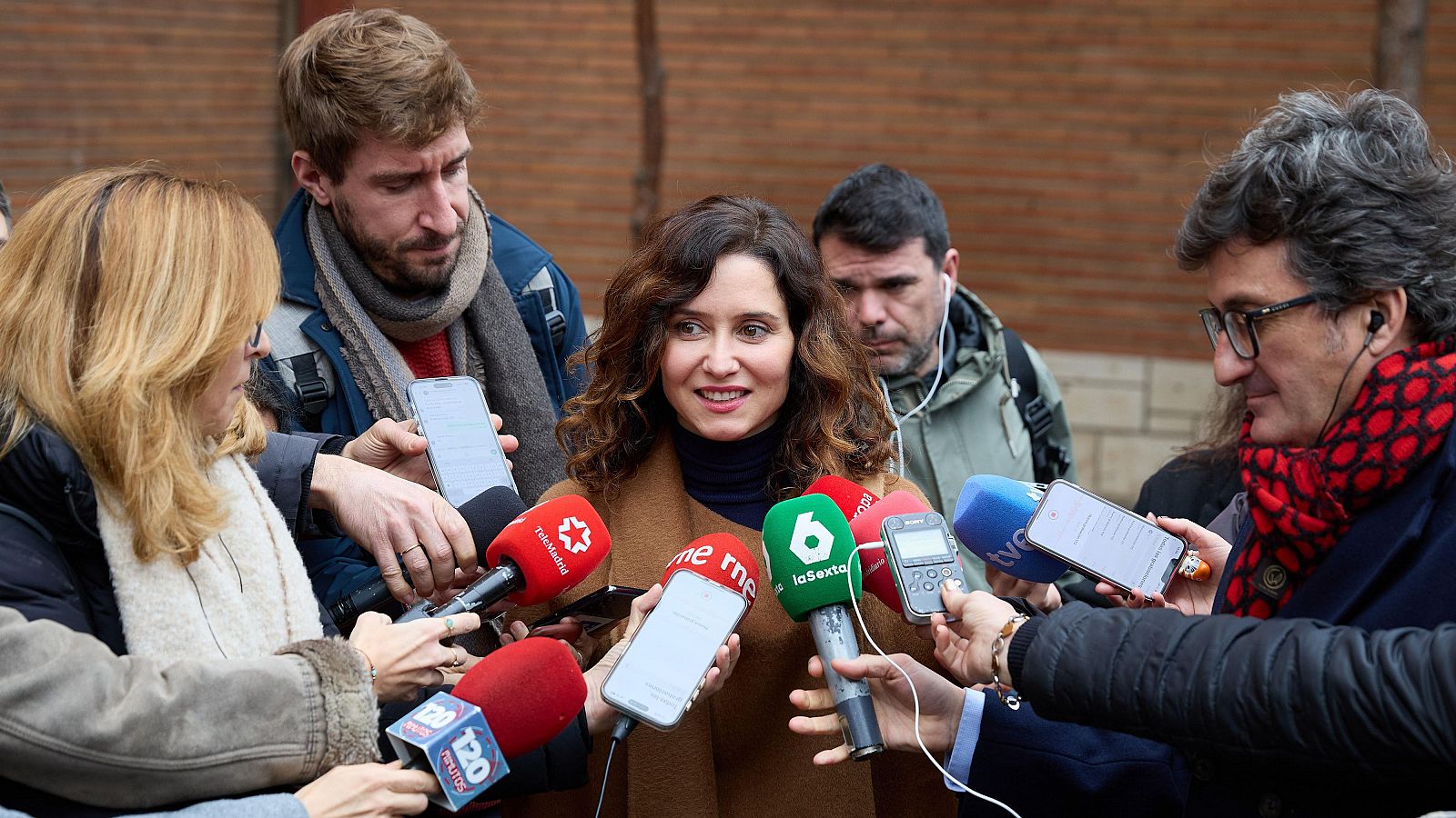 La presidenta de la Comunidad de Madrid atiende a los medios, rodeada de periodistas con micrófonos y grabadoras en un acto público exterior.