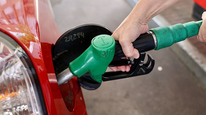 Mano sujetando manguera verde de gasolina mientras reposta un coche rojo.  Imagen cercana que resalta la acción de repostar combustible.