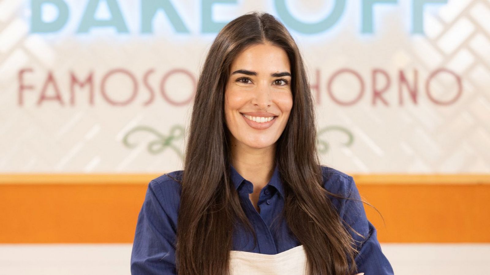 Lidia Torrent, en 'Bake Off Famosos', sonríe a cámara con camisa azul marino y delantal beige.  Se observa el logo del programa al fondo.