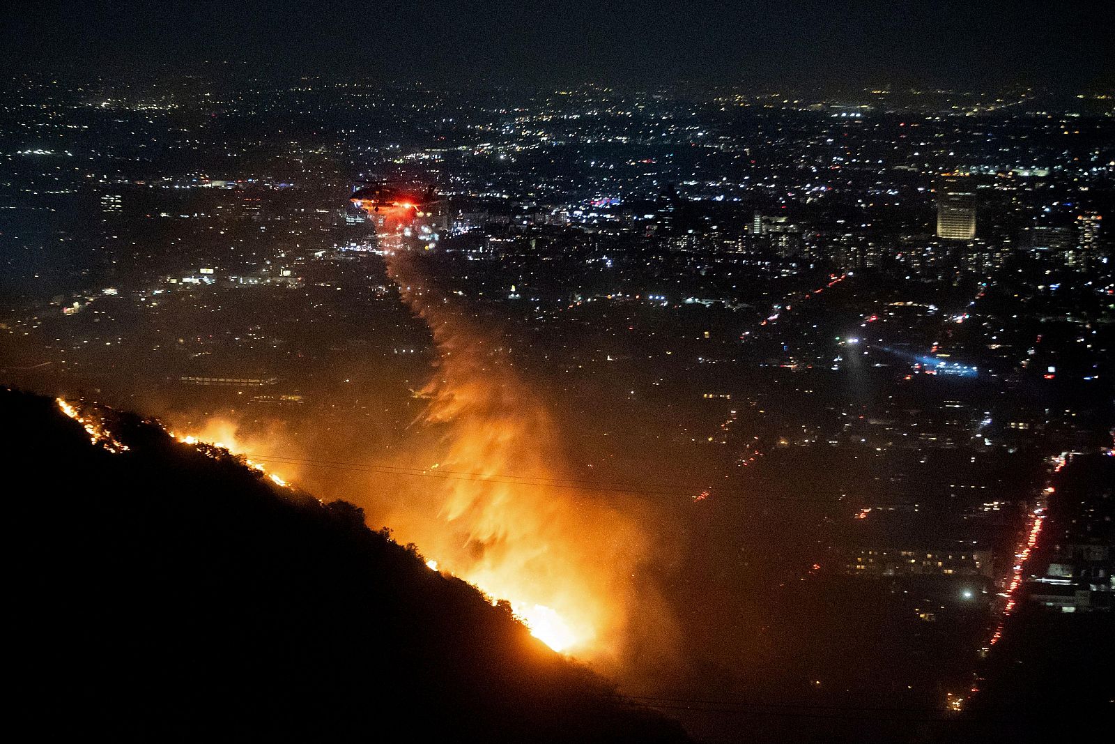 Helicóptero apaga incendio forestal nocturno con grandes llamas y humo en una ladera.  Ciudad iluminada al fondo.