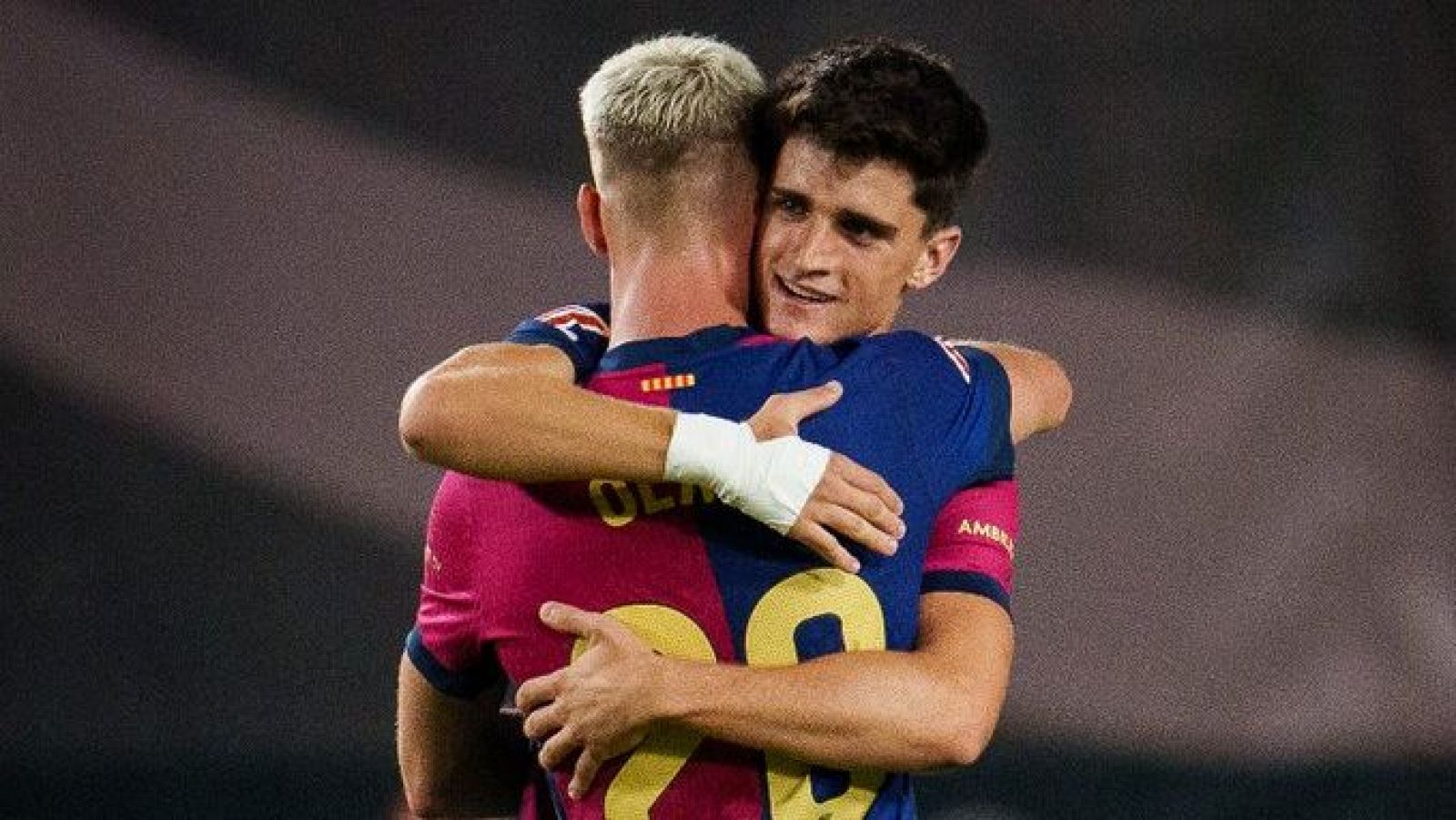 Abrazo entre dos jugadores de fútbol, uno con camiseta granate y dorsal 20, el otro con camiseta azul marino.  Muestran alegría y camaradería.