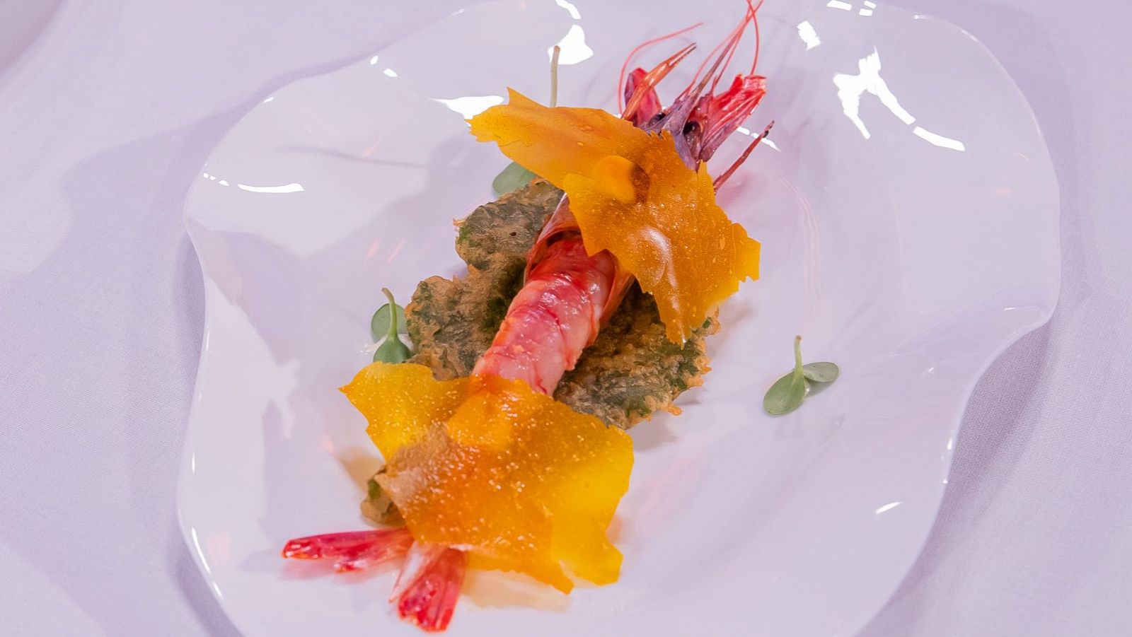 Gamba roja a la plancha sobre tempura crujiente, decorada con láminas naranjas y hojas verdes. Presentación cuidada en plato blanco.