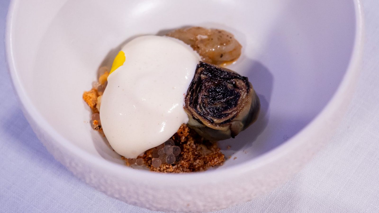 Alcachofa confitada oscura con tartar de langostinos claro, cubiertos por espuma blanca y detalles marrones y amarillos. Presentación cuidada.