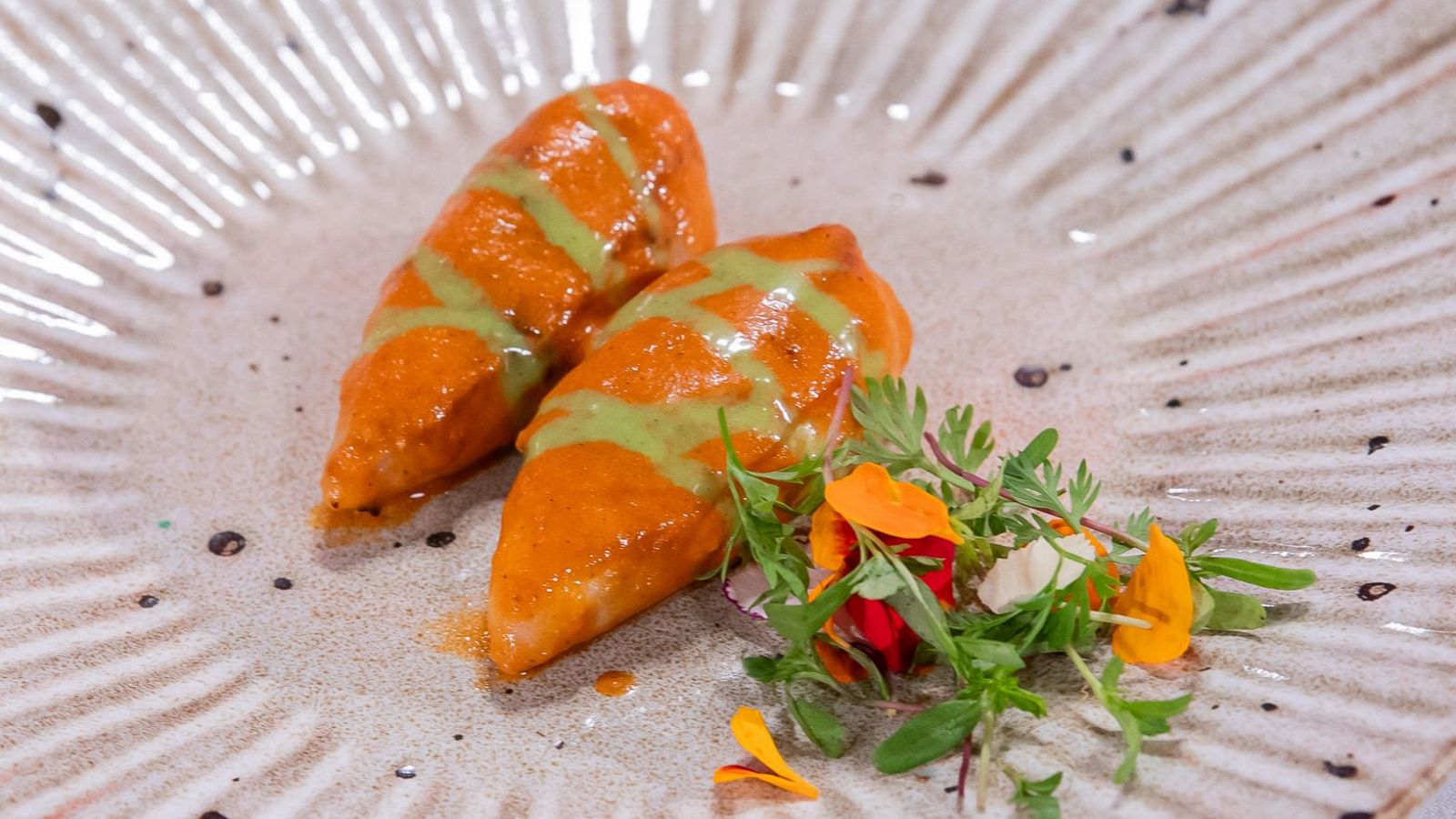 Plato de cerámica con dos chipirones rellenos, salsa rojiza y verde, guarnición de hojas verdes, flores y queso.