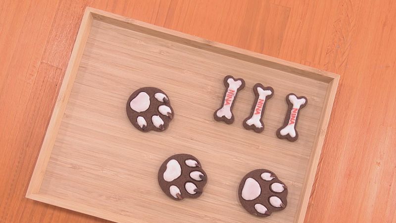 Galletas de cacao con forma de hueso y huella canina, algunas con el nombre "NINA" en naranja, presentadas en bandeja de madera.