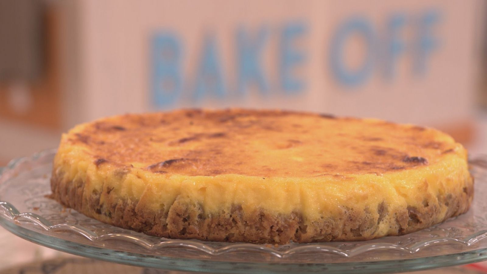 Tarta de queso y membrillo dorada, con base oscura, sobre bandeja. Fondo difuminado con texto "BAKE OFF".