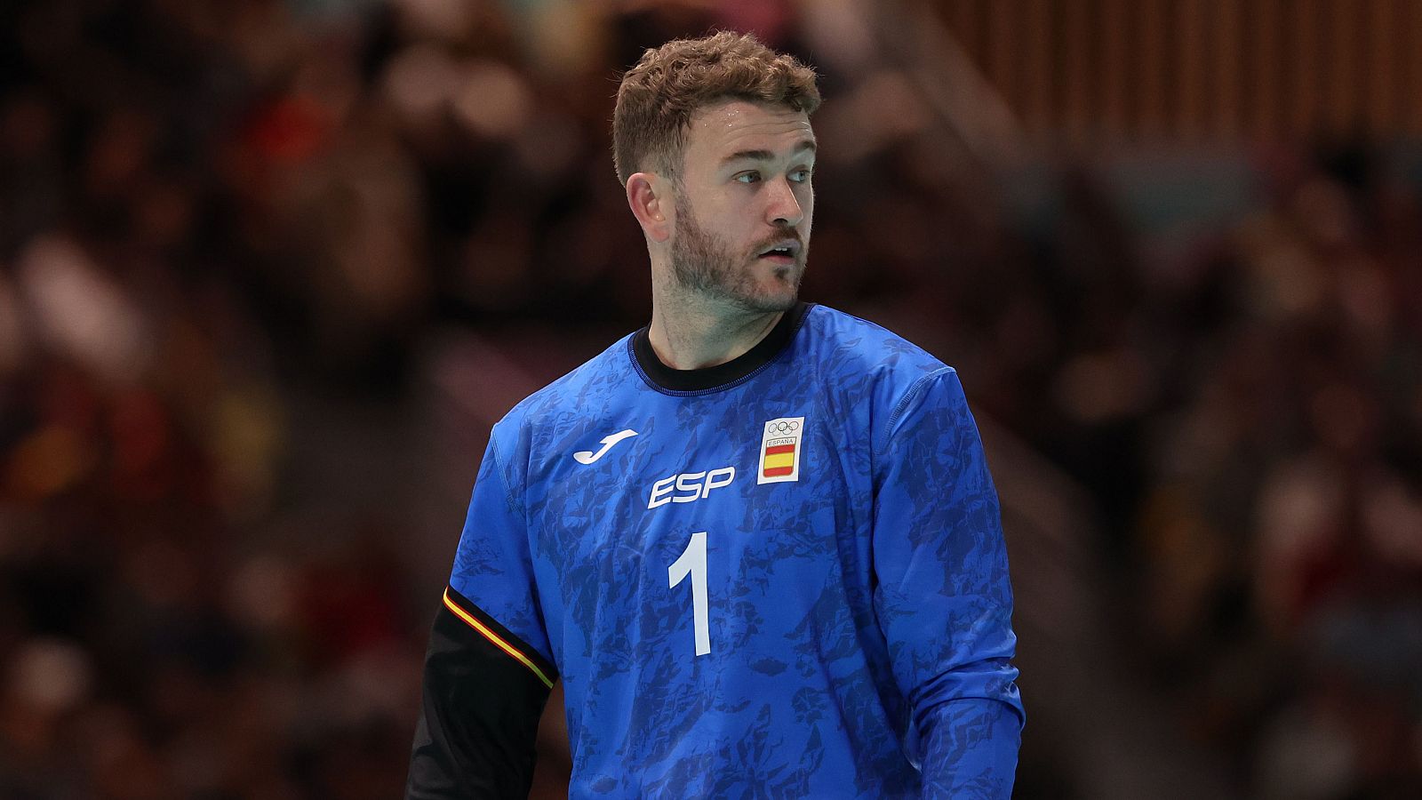 Portero español de balonmano, camiseta azul con el número 1 y logo Joma, expresión concentrada. Fondo con público desenfocado.