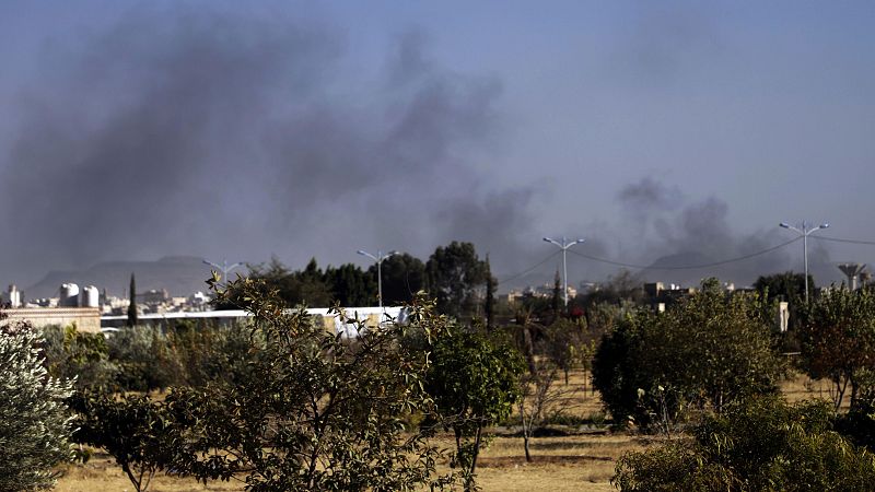Imagen de un paisaje devastado por bombardeos: humo negro, edificios dañados y un terreno árido.  Se aprecian postes eléctricos y cables.