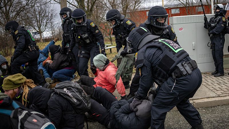Policías antidisturbios controlan una manifestación, algunos manifestantes sentados en el suelo son retirados.  Un agente sujeta a una persona con chaqueta rosa.
