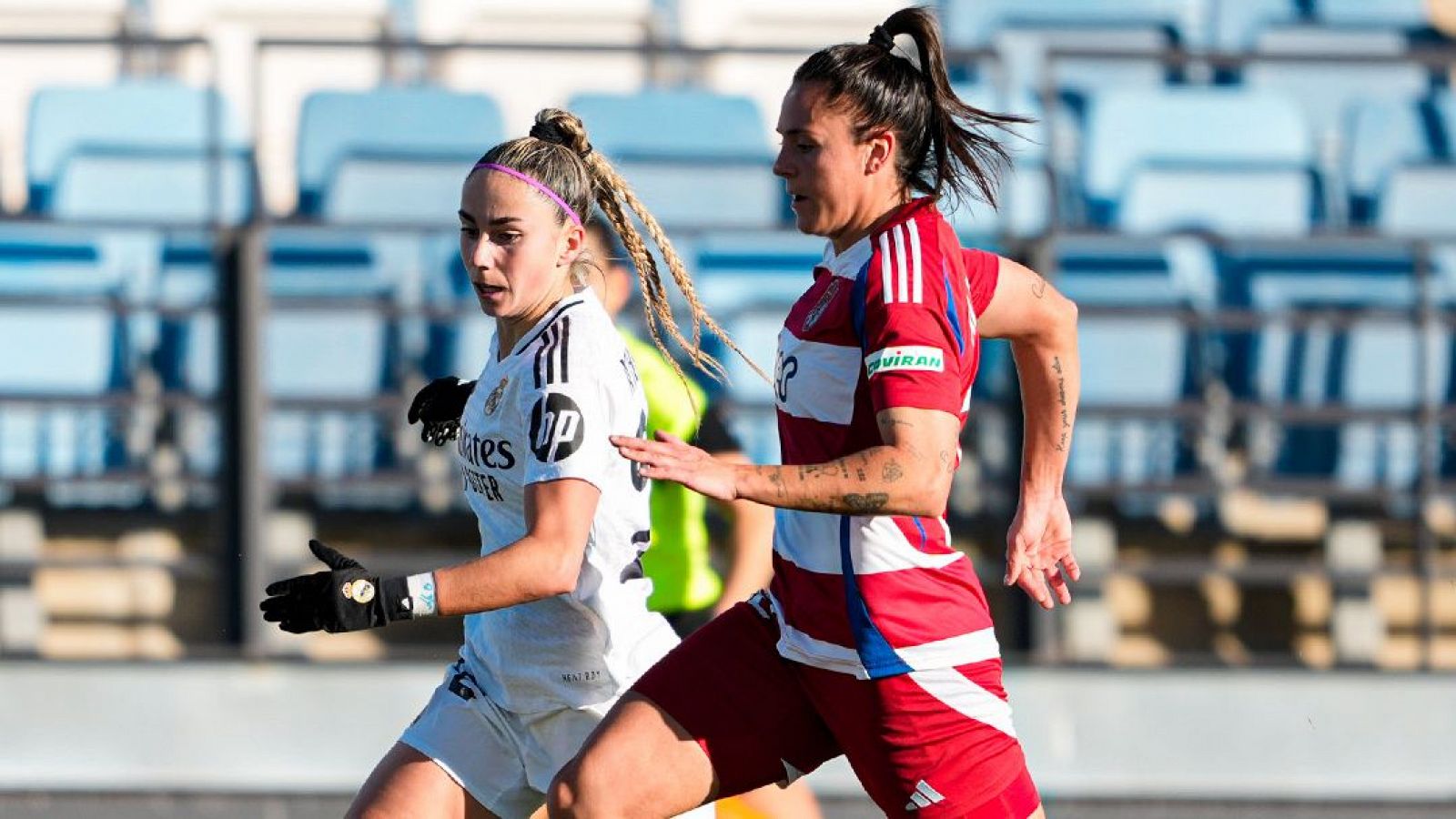 Partido de fútbol femenino: dos jugadoras, una con camiseta blanca y otra con camiseta roja y blanca, corren tras el balón en una escena llena de acción.