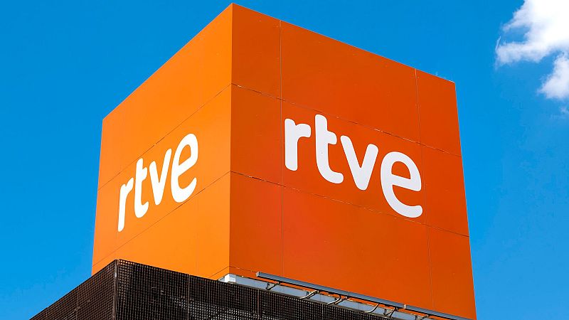 Rótulo cúbico naranja con logotipo RTVE en blanco, situado en la parte superior de un edificio de color marrón oscuro con estructura metálica visible. Cielo azul de fondo.