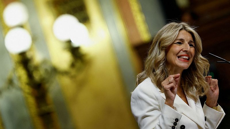 Imagen de Yolanda Díaz, con americana blanca y pelo rubio ondulado, hablando o gesticulando en un entorno institucional.