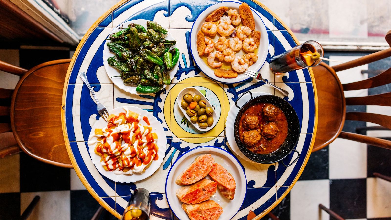 Mesa con tapas españolas (pimientos, gambas, patatas bravas, albóndigas) servidas en platos pequeños sobre un mantel de azulejos. Dos vasos y sillas de madera completan la escena.