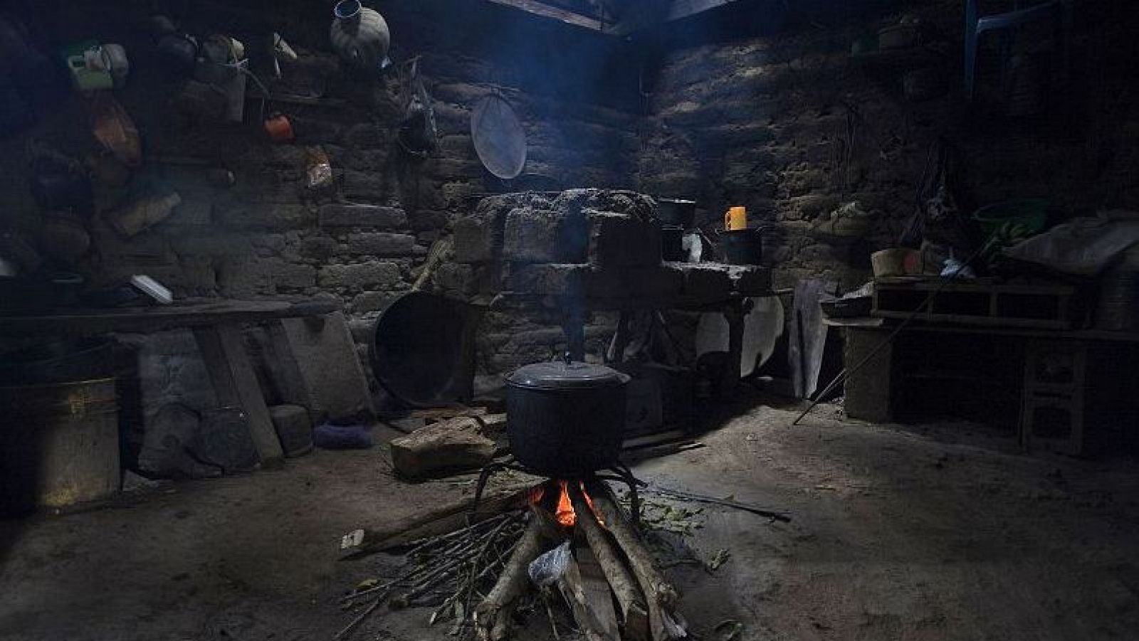 Cocina rústica guatemalteca con fuego de leña, ollas, sartenes y utensilios colgados en paredes de piedra y ladrillo. Ambiente oscuro y con poca ventilación.