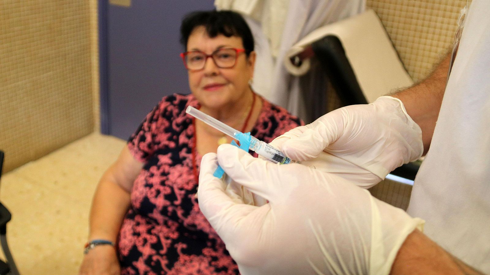 Paciente con gafas y blusa floral observa a un sanitario con guantes preparar una inyección en una consulta médica.  Vacunación contra la gripe.