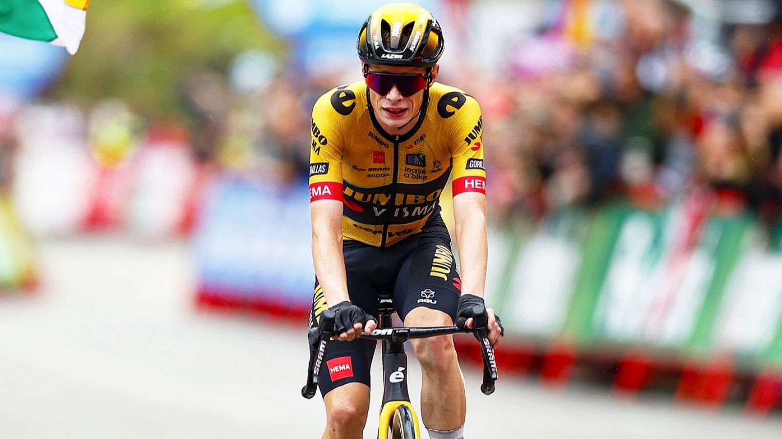 Ciclista con maillot amarillo, gafas de sol y casco, agarrando el manillar de su bicicleta negra durante una carrera. Fondo con público y publicidad.