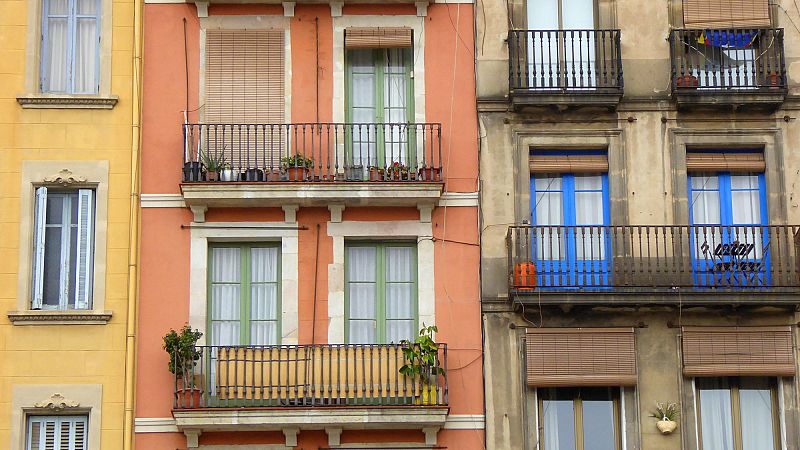 Fachadas de tres edificios: dos anaranjados/rojizos, uno gris; todos con balcones, ventanas azules en el central, plantas en macetas.