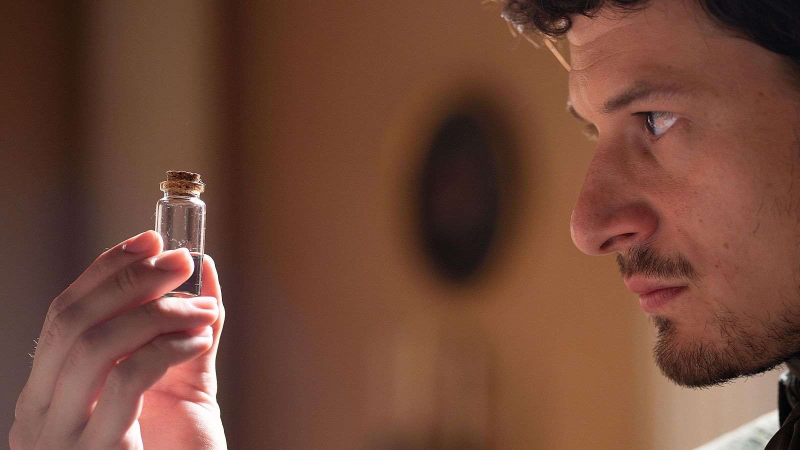 Hombre con cabello oscuro y rizado, expresión seria, sostiene un frasco con líquido oscuro.  Imagen de una serie, posible preparación para una misión.