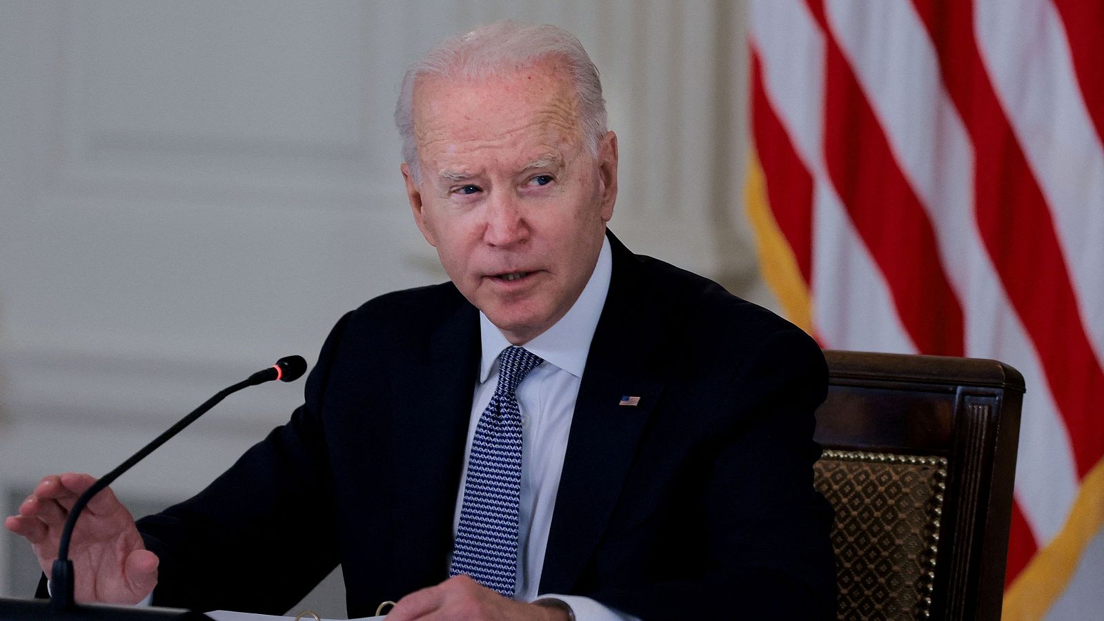 El presidente Biden, en traje oscuro y corbata con lunares, parece dirigirse a alguien en una reunión formal con una bandera estadounidense de fondo.