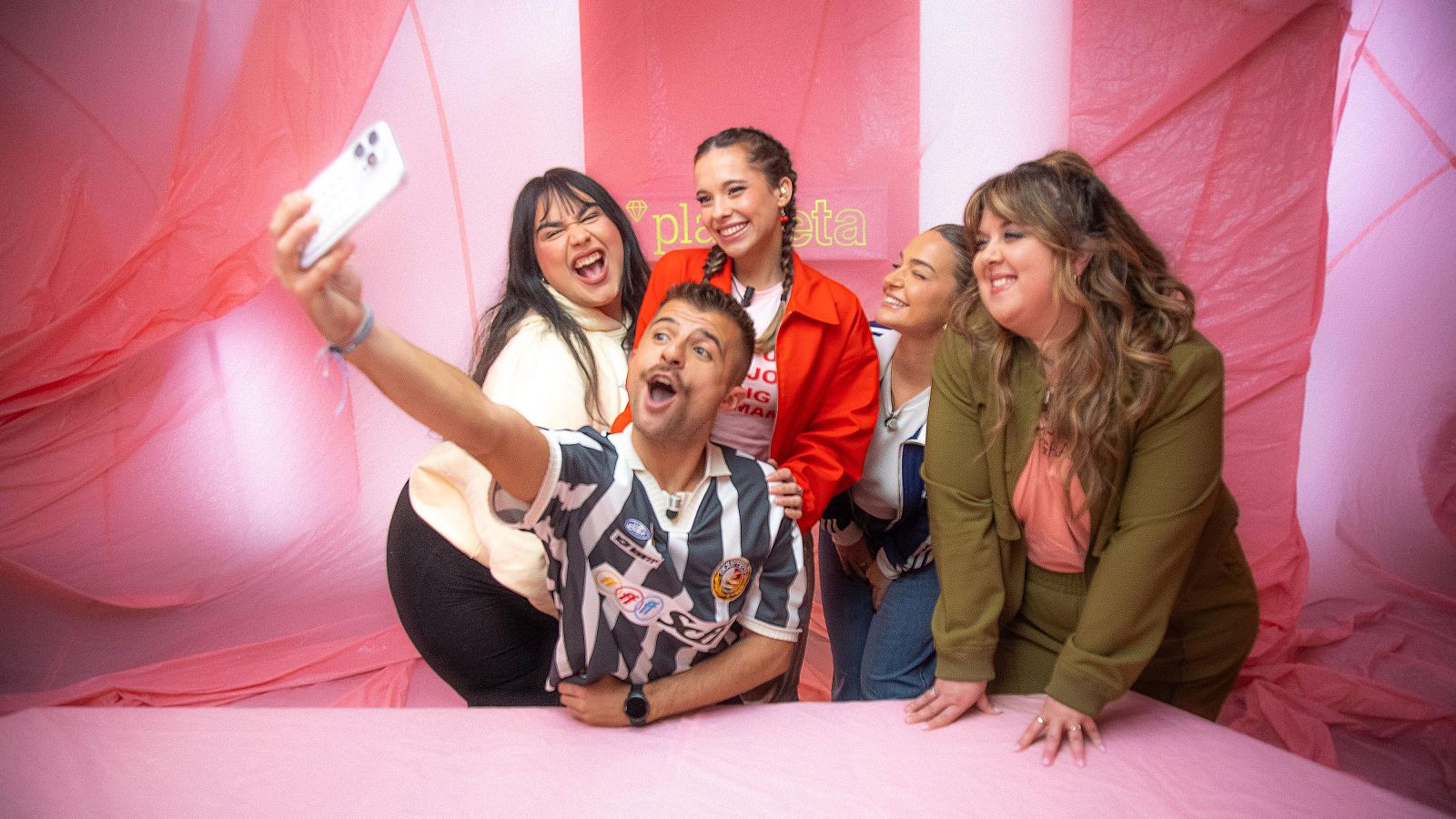 Selfie grupal en un escenario rosa.  Se ven varias personas con ropa variada: camiseta deportiva, chaqueta roja, prendas blancas y verde oliva. Ambiente juvenil.