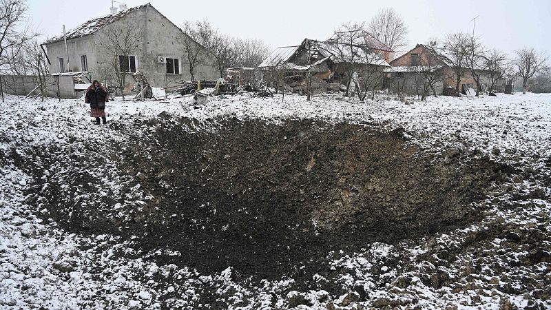 Cráter en terreno nevado junto a dos casas, una dañada. Mujer observa la escena de devastación. Impacto de la guerra.