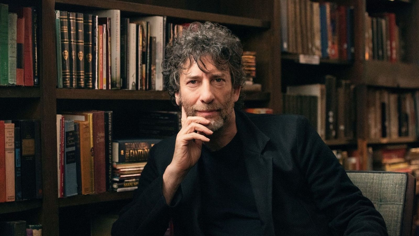 Neil Gaiman, sentado, mira a cámara con expresión serena;  una estantería llena de libros forma el fondo de la imagen.