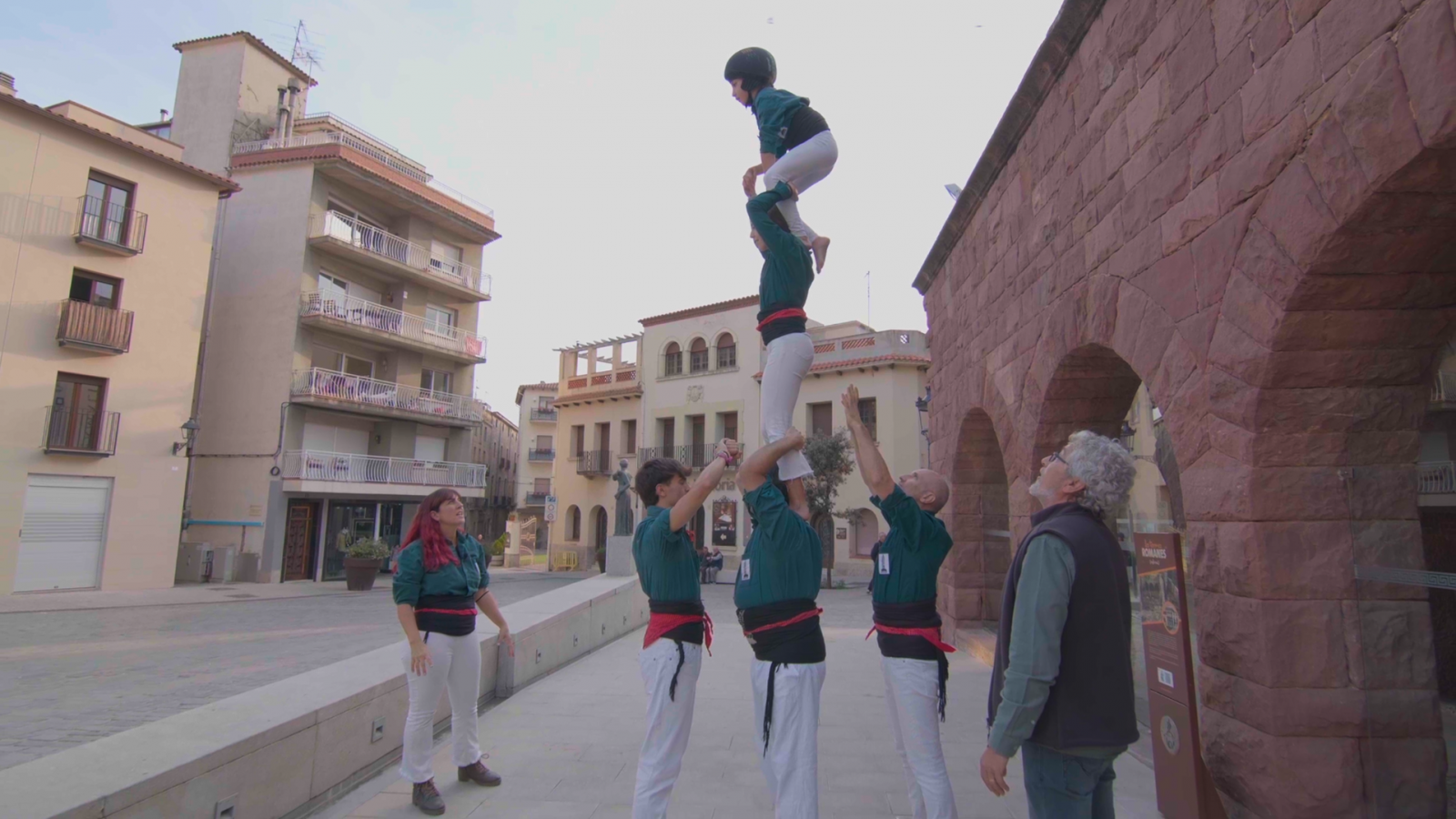 Construcción de un castell con una niña en la cima usando casco.  Participantes con ropa variada, incluyendo camisas verde oscuro y pantalones blancos.  Grabación o ensayo de un programa de TV.