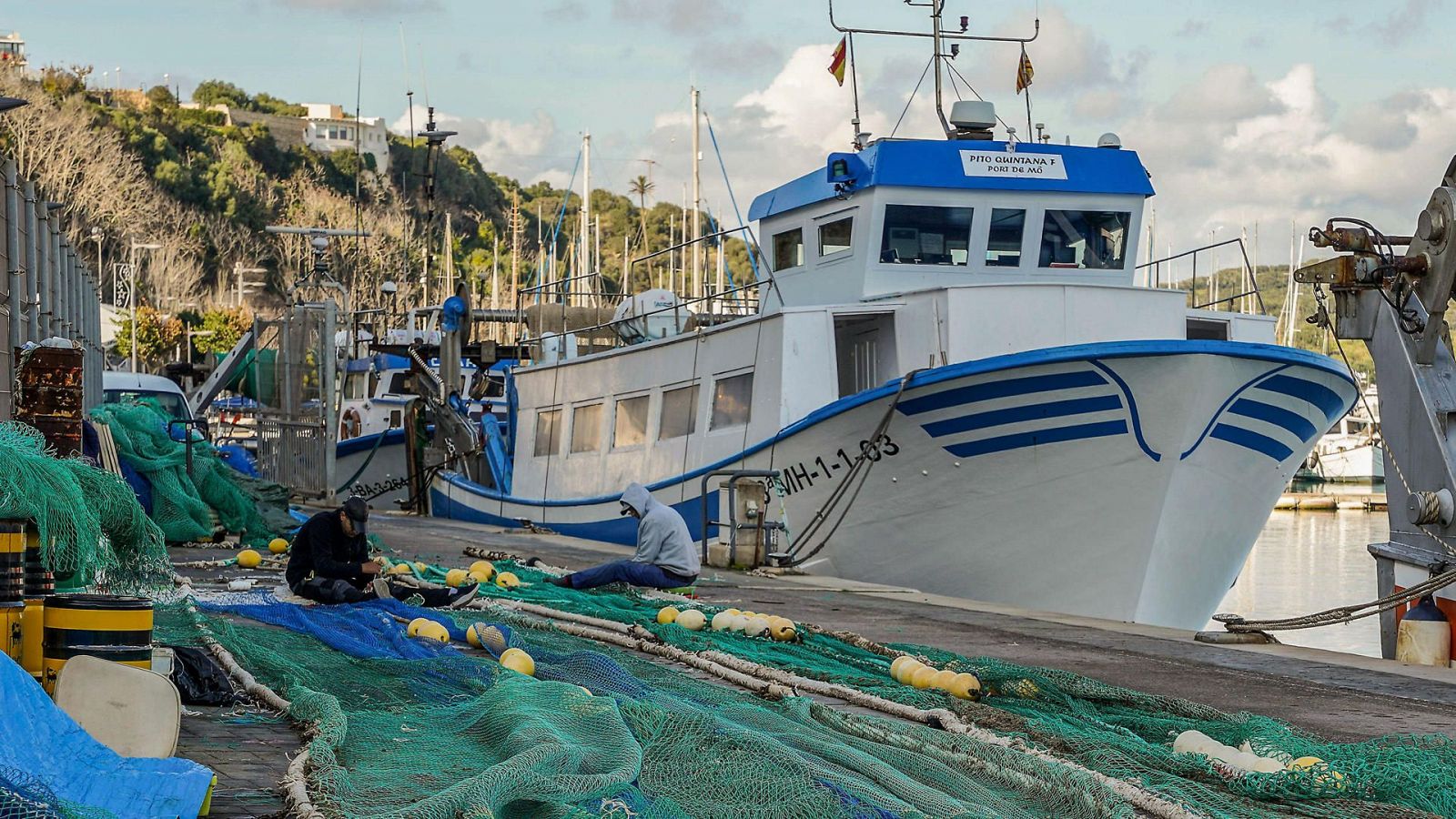 Flota pesquera en puerto, inactiva por huelga. Barco blanco con franjas azules y matrícula visible. Pescadores en el muelle junto a redes.