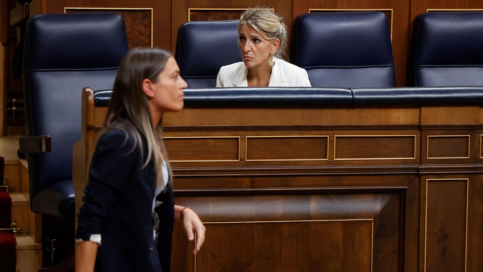 Yolanda Díaz, sentada, y Miriam Nogueras, de pie, en el hemiciclo del Congreso durante una sesión parlamentaria. Díaz viste chaqueta blanca y Nogueras americana oscura.