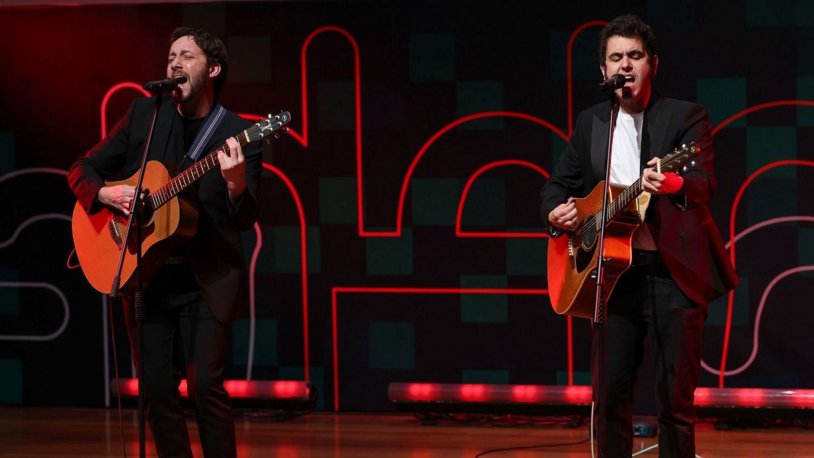 Actuación musical de dos hombres con guitarras acústicas en un escenario con iluminación roja y verde. Visten trajes oscuros y camisas claras.