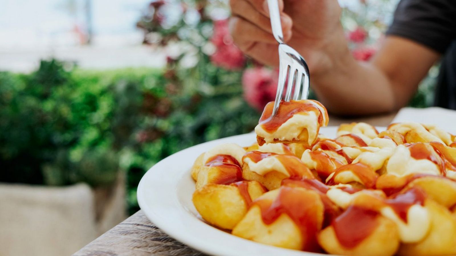 Patatas bravas con salsa roja y blanca, pinchadas con un tenedor. Primer plano sobre mesa de madera con fondo desenfocado de vegetación.