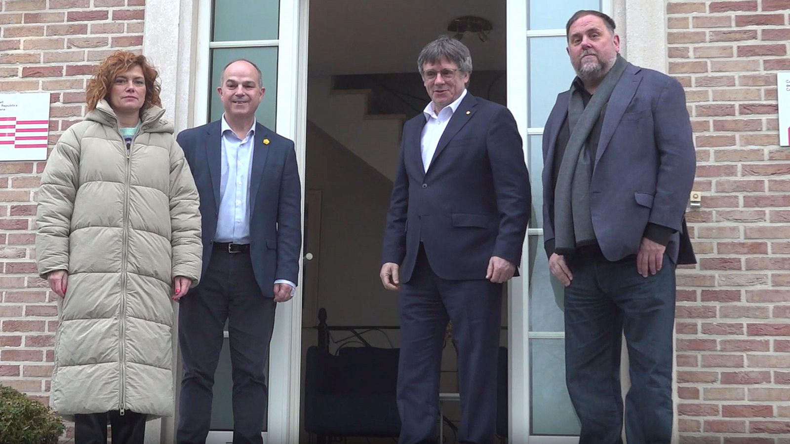 Reunión de cuatro figuras políticas catalanas frente a un edificio de ladrillo en Waterloo. Se aprecian diferentes estilos de vestimenta: abrigos, trajes y vaqueros.