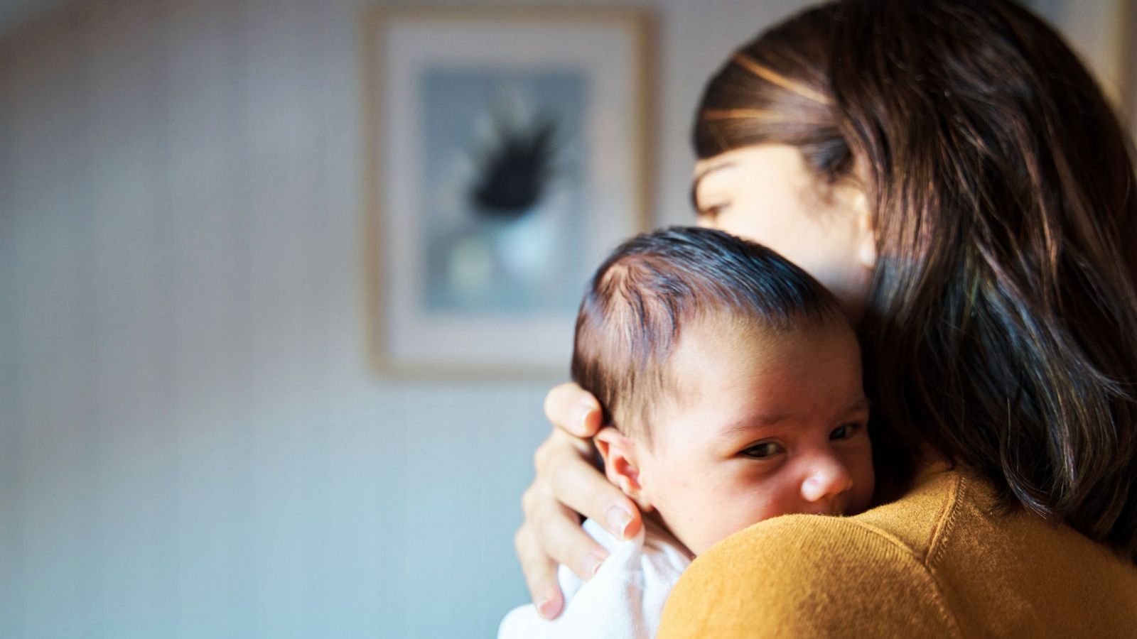 Madre con bebé de 1-3 meses. La madre, de espaldas, viste de mostaza; el bebé, de blanco, mira a la cámara. Fondo desenfocado con pared clara y cuadro.