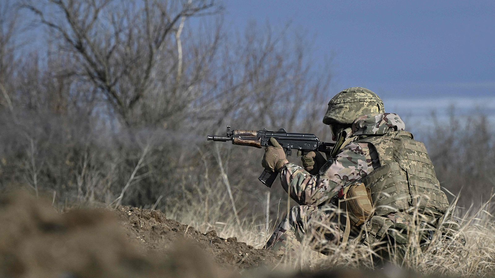 Soldado en entrenamiento táctico, agachado y apuntando con un rifle que acaba de disparar.  Se observa humo y un terreno árido.