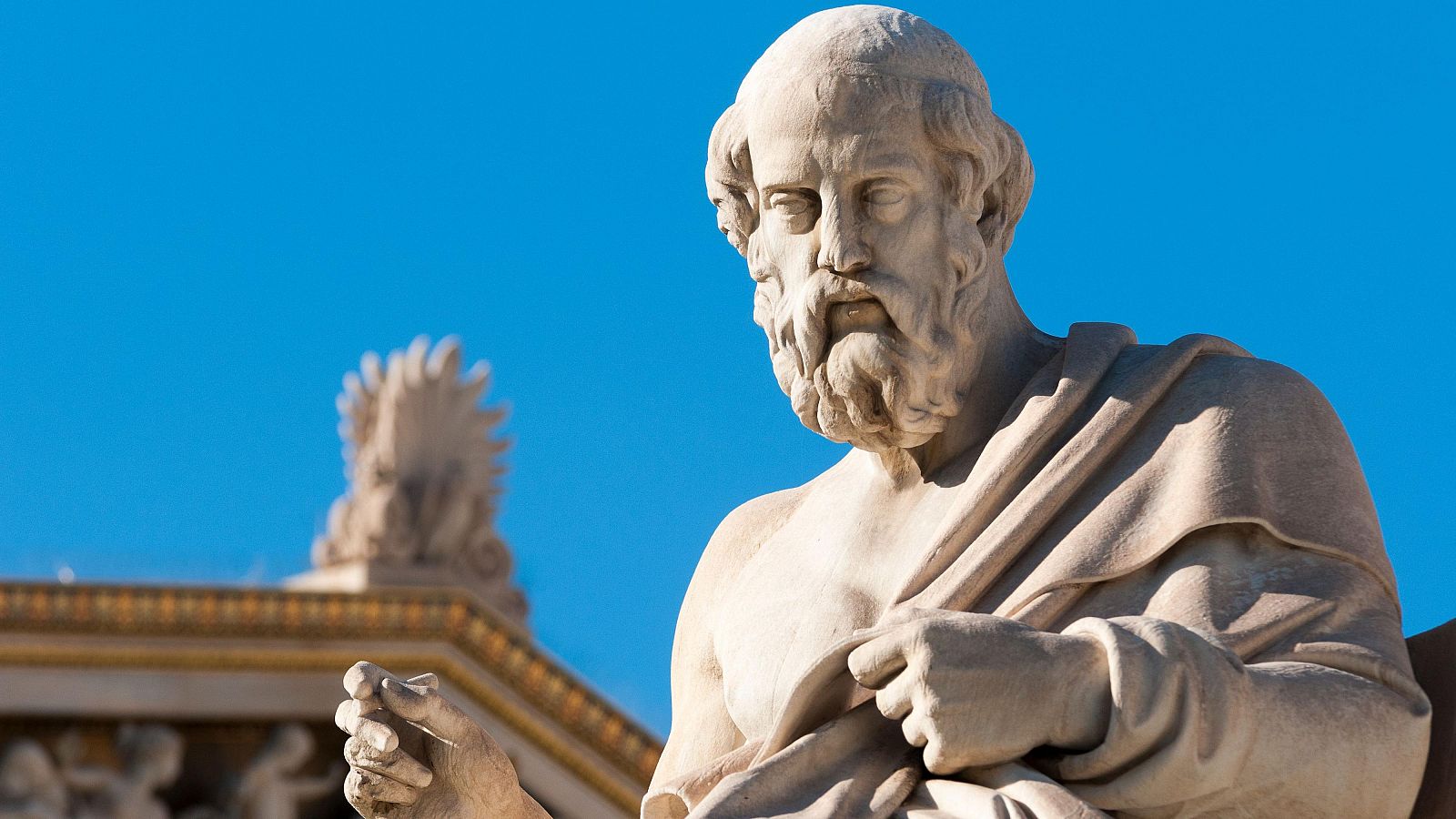 Estatua de mármol blanco de Platón, sentado con barba y toga, gesticulando. Fondo con cielo azul y detalles dorados de un edificio.