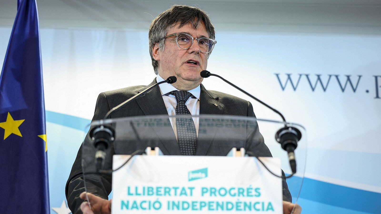 Puigdemont, con traje oscuro, habla desde un atril con el lema "LLIBERTAT PROGRÉS NACIÓ INDEPENDÈNCIA", con una bandera europea visible.  Fotografía: EFE/Pablo Garrigós.