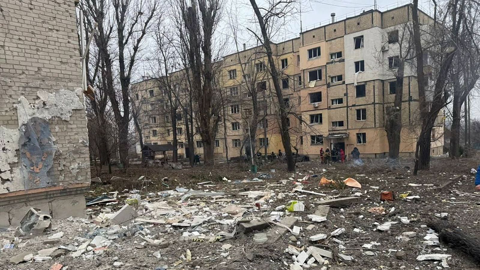 Daños considerables en un edificio tras un bombardeo. Escombros, personas (posiblemente servicios de emergencia) y árboles sin hojas en la escena.