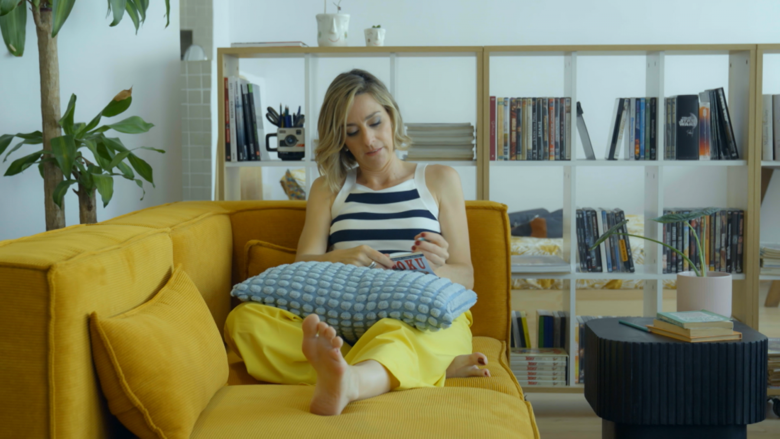 Mujer rubia sentada en sofá mostaza, leyendo o tejiendo, rodeada de libros y plantas. Ambiente relajado y hogareño.