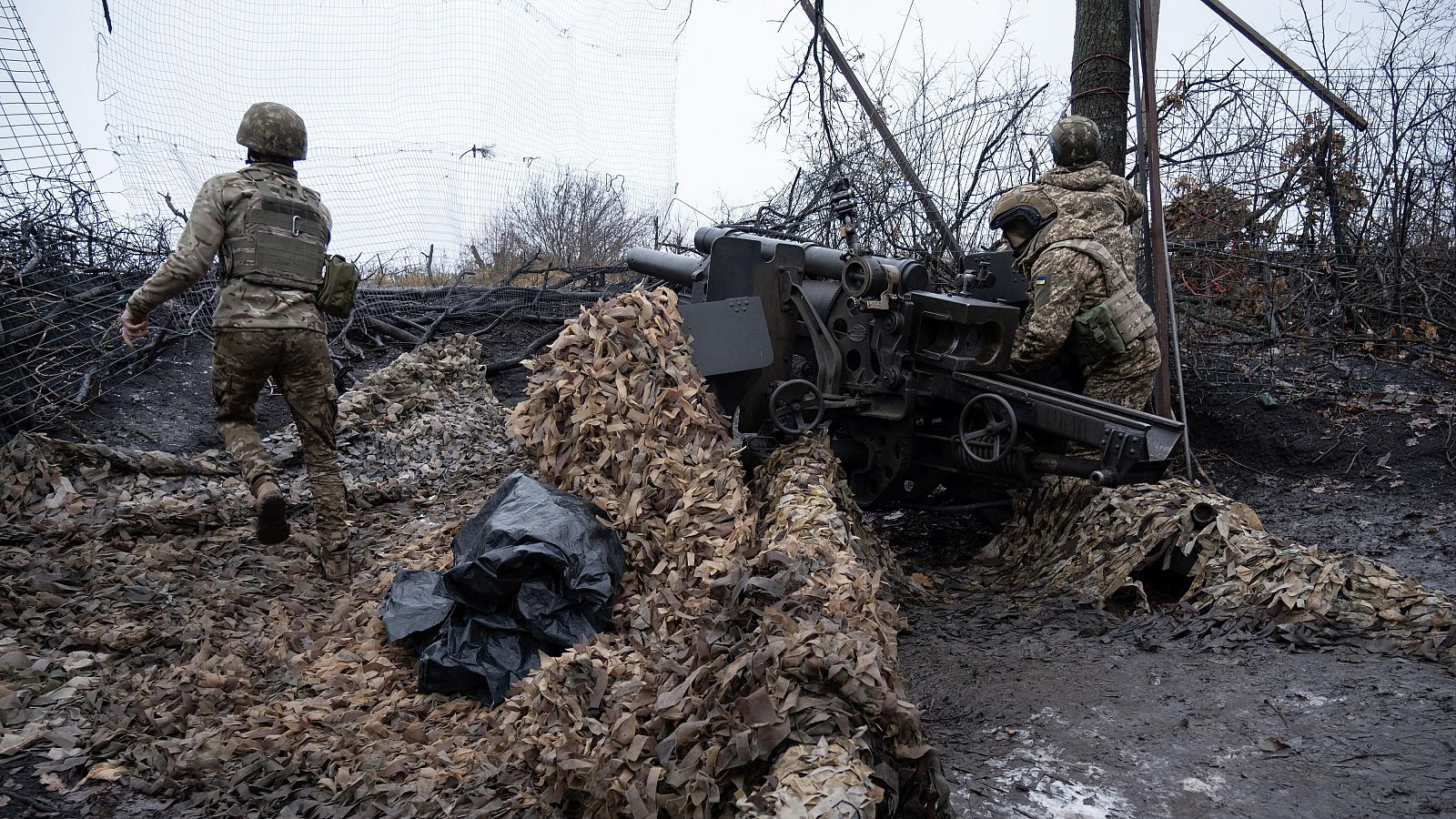 Obús camuflado operado por dos soldados ucranianos en un terreno embarrado con vegetación quemada. Un tercer soldado se aleja.