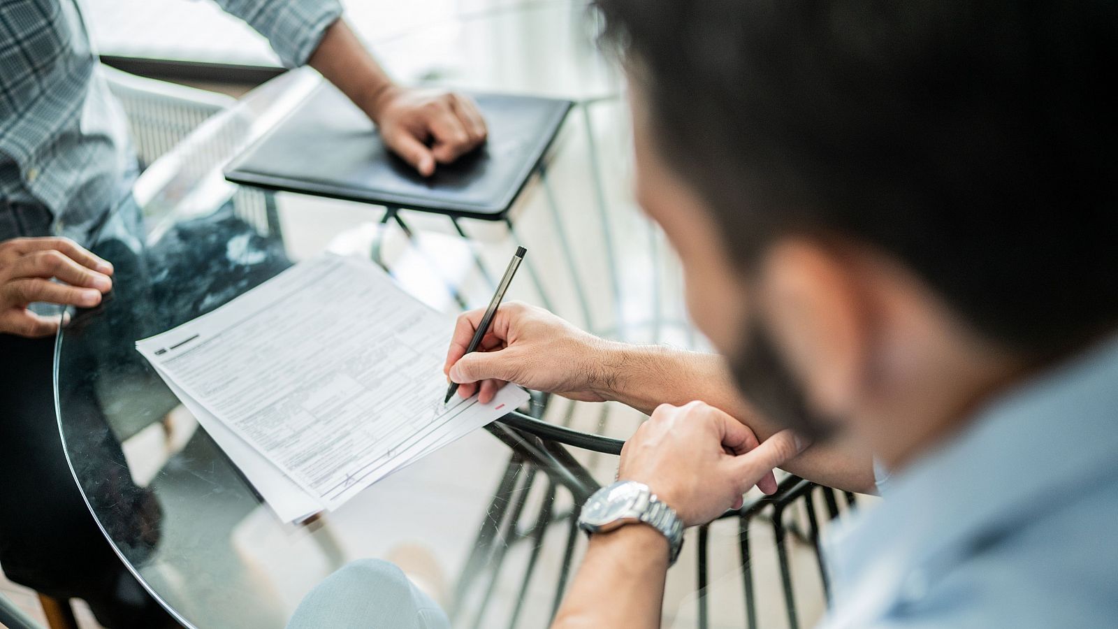 Firma de un documento en una mesa de cristal. Una persona con camisa azul claro firma, mientras otra con camisa a cuadros observa.