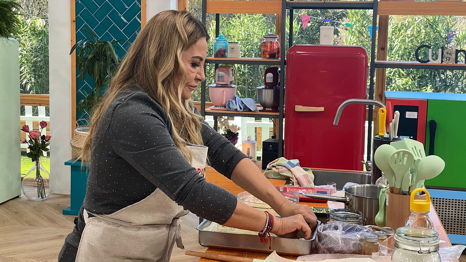 Presentadora con delantal beige manipulando una bandeja metálica en una cocina moderna con electrodomésticos coloridos y detalles decorativos.