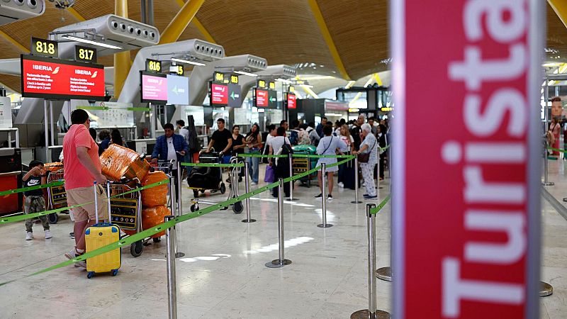 Larga cola de pasajeros en un aeropuerto con maletas, mostradores de facturación con pantallas mostrando 'Iberia' y números de vuelo, y un cartel borroso con la palabra 'Turista'.