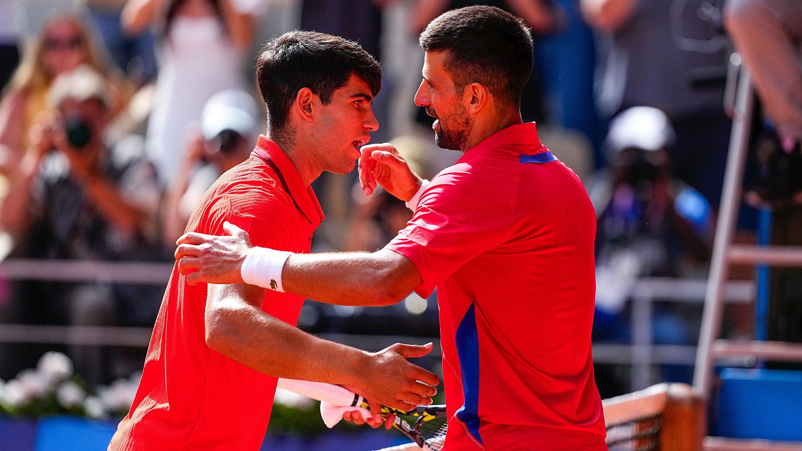 Dos tenistas, vestidos con camisetas rojas, se abrazan en una pista tras una final.  La escena muestra deportividad y respeto.