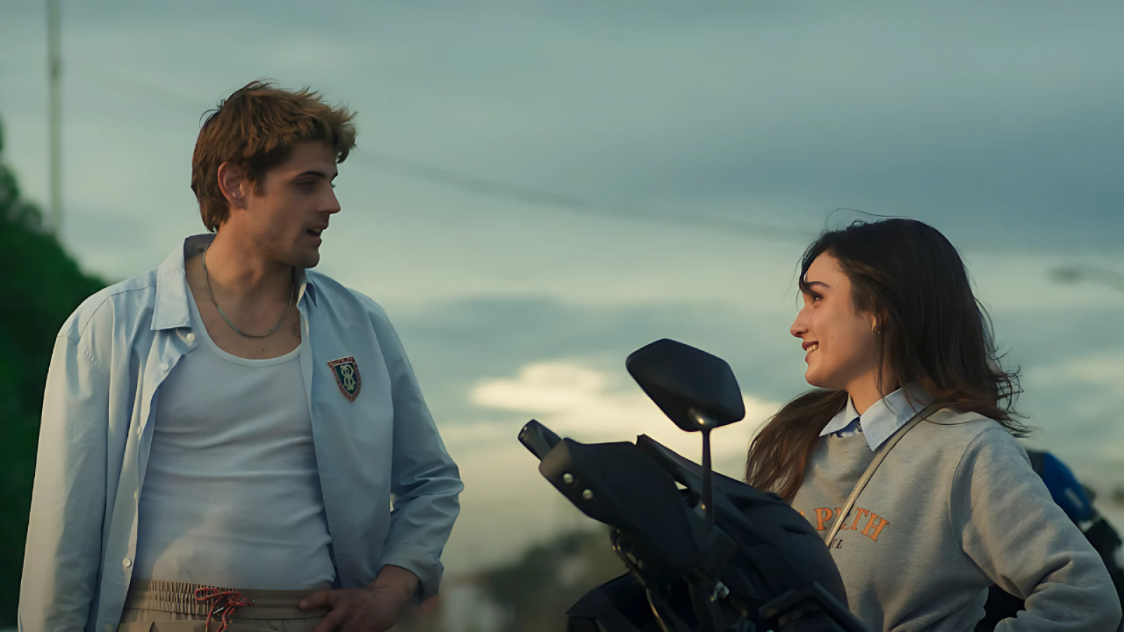 Dos jóvenes, con ropa informal, sonríen en una escena al aire libre, posiblemente al atardecer.  Se observa parte de una motocicleta.