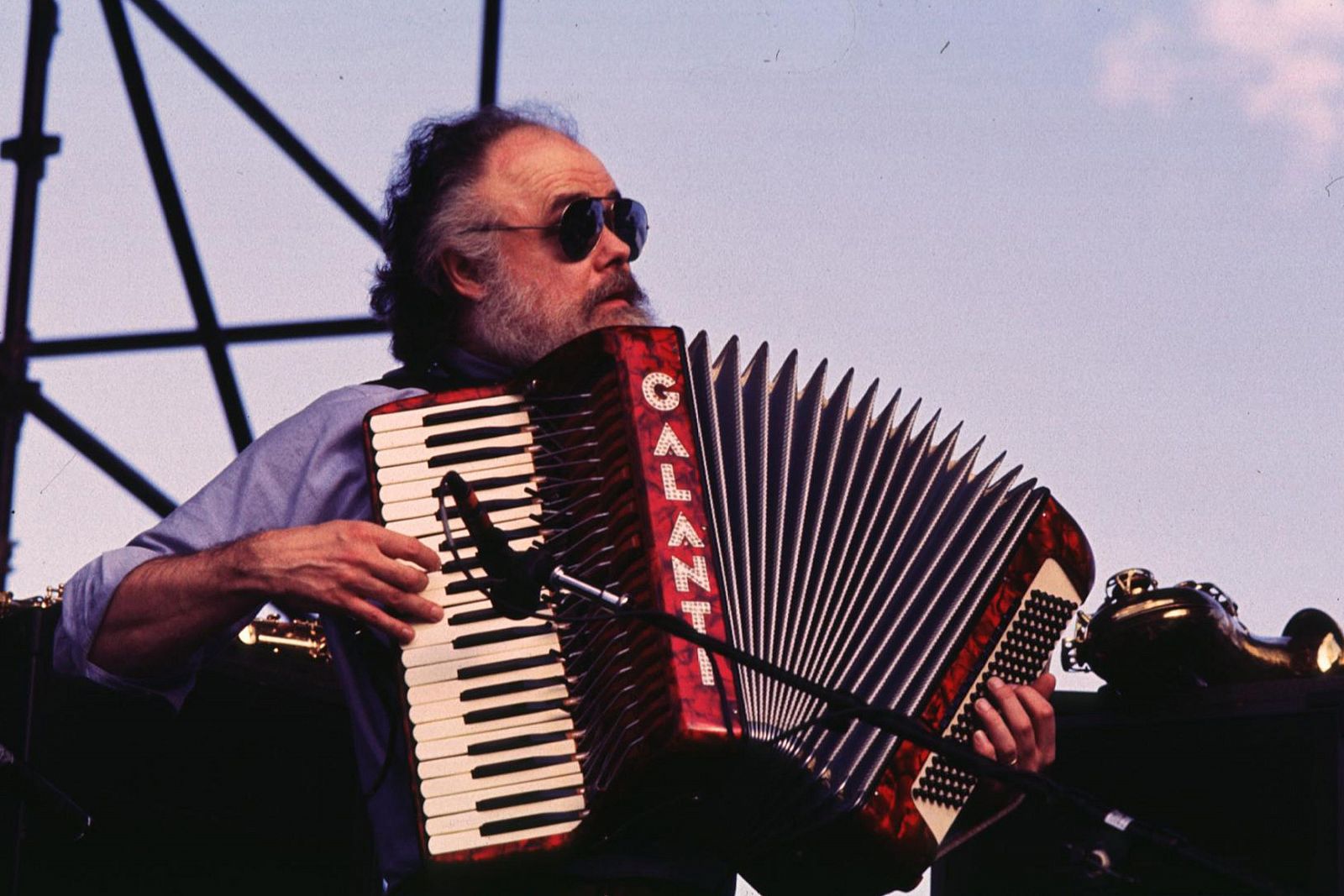 Músico con barba y gafas de sol tocando un acordeón en un escenario exterior.  Se aprecia un instrumento de viento en la esquina inferior derecha.