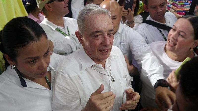 Álvaro Uribe en una concentración, rodeado de gente; dos mujeres cercanas a él, una a cada lado.  Un hombre con gafete también está presente.