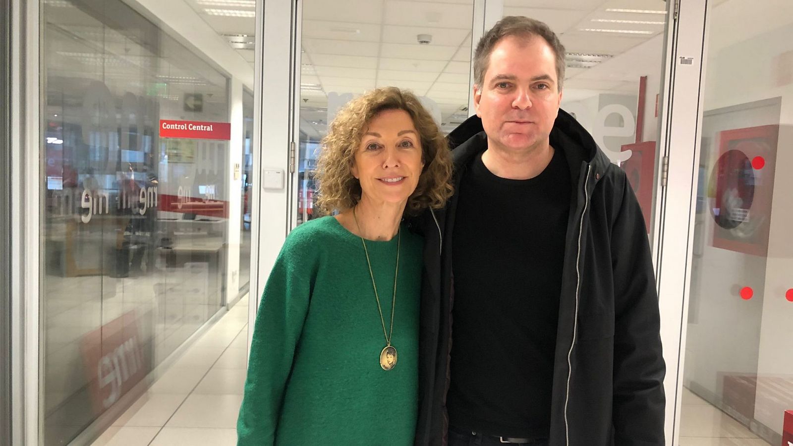 Foto de dos personas (mujer con jersey verde, hombre con chaqueta negra) en un pasillo con logo de RNE.  Posiblemente en las instalaciones de Radio Nacional de España.