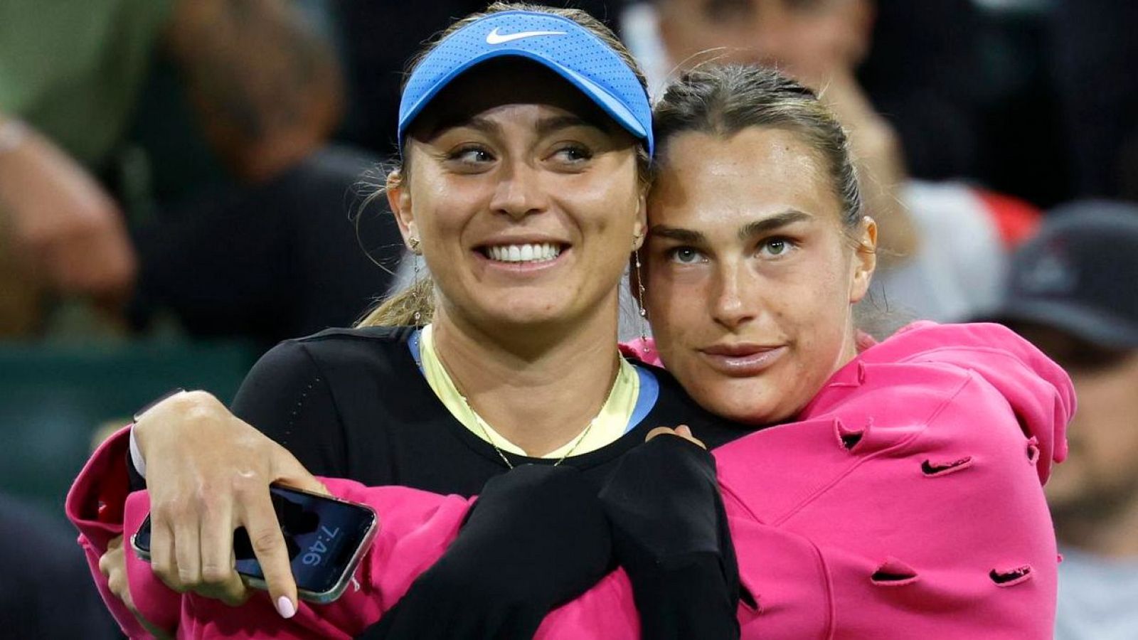 Dos tenistas, una con visera azul y otra con sudadera rosa, se abrazan y sonríen tras un partido.  Una de ellas muestra un móvil con la hora.