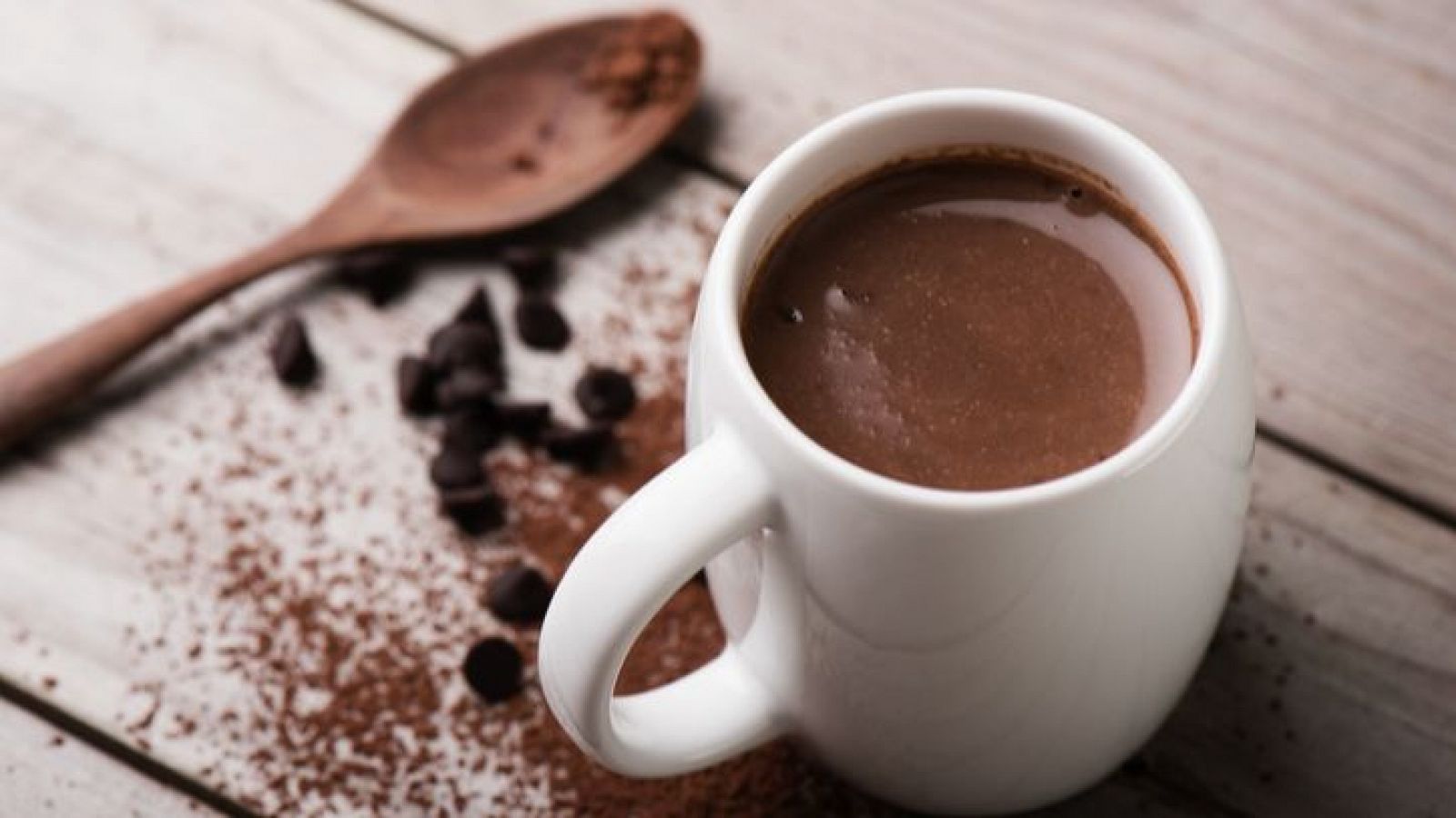 Chocolate caliente espeso y humeante en taza blanca, sobre mesa de madera con chips de chocolate y cacao en polvo, junto a una cuchara de madera.